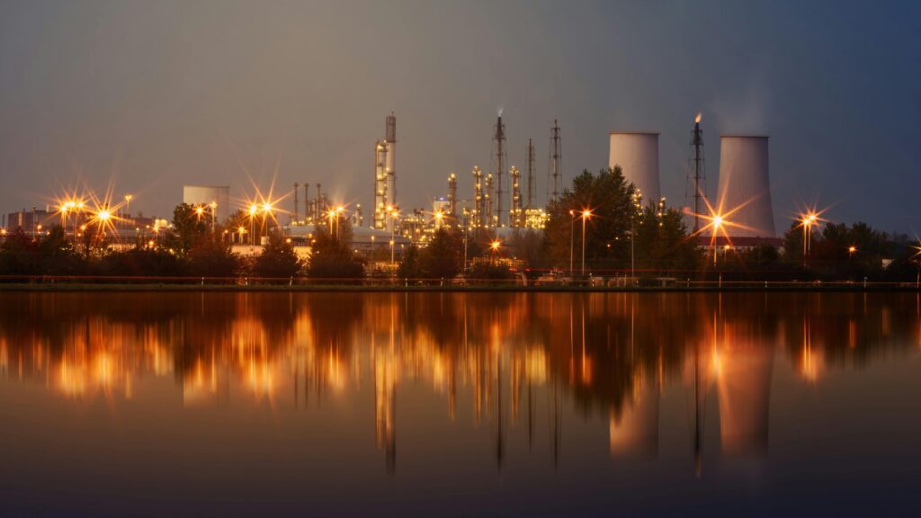 Projects Industrial plant with reflection in water at twilight, showing smoke stacks and lights.